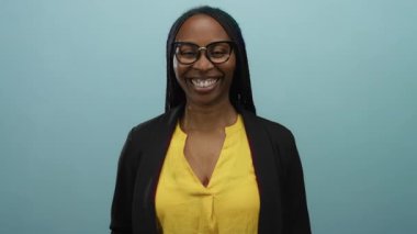 Sarı bir bluz içinde gülümseyen gözlüklü siyah kadın mavi bir duvarın arkasında duruyor..