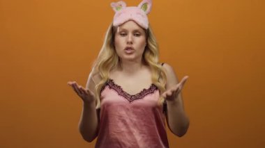 Kadın gece elbisesi giyiyor, sarı saçlı, turuncu arka planda, gece elbisesiyle göz maskesiyle duygularını ifade ediyor, dinleyicileri canlı bir şekilde kendine çekiyor..