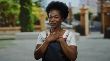 Kadın açık havada göğsünde ellerle gülümsüyor, önlük giyiyor, bir şehir caddesi düzeninde, kentin ortasında bir memnuniyet ve minnettarlık duygusu somutlaştırıyor..