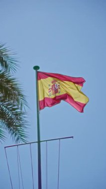 İspanya bayrağı açık mavi gökyüzüne karşı dalgalanırken palmiye yaprakları nazikçe sallanırken, İspanya 'nın canlı kültürünü ve ılık iklimini simgeliyor..