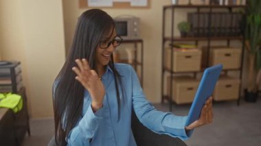 Kadın, ofis ortamında kitap ve ofis dekorlarıyla çevrili bir tablet kullanarak profesyonel ama gelişigüzel bir etkileşim kurarken gülümsüyor..