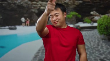 Tatil köyü havuzundaki Asyalı adam açık hava otel ortamında neşeli jestler yapıyor. Kendine güveni ve çekici tavrını doğal çevresi olan günlük kıyafetleriyle ifade ediyor..