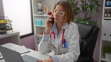 Doktor konuşuyor, klinikte çalışan sarışın, beyaz tenli, gözlüklü, kapalı alanda çalışan, bilgisayarlı bir bayan..