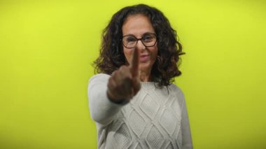 Orta yaşlı, kıvırcık saçlı ve gözlüklü İspanyol kadın gülümsüyor. Sarı arka planda kalp hareketi yapıyor. Sıcaklık ve pozitiflik taşıyor..