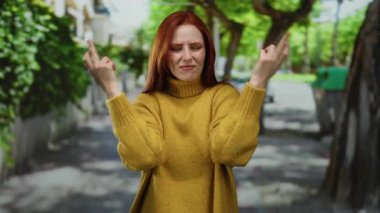 Şehir caddesinde, sarı süveter giyen, ağaçlar ve binalar arka planda sıralanırken kızıl saçlı, hayal kırıklığına uğramış jestler yapan bir kadın..