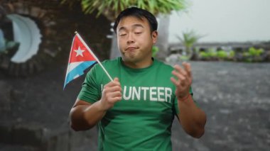 Şehir ortamında Küba bayrağı taşıyan genç bir adam, yeşil gönüllü tişört giyiyor, bir şehir ortamında uluslararası kültür veya gönüllü bağlantı öneriyor..