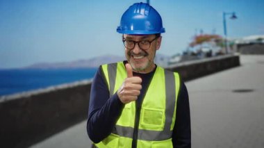 Kask ve güvenlik yeleği giyen İspanyol bir adam açık gökyüzünün altında deniz kenarında bir gezinti için onay veriyor..