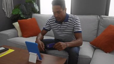 Çizgili tişörtlü genç adam, modern oturma odasında rahat kanepe ve bitkiler içeren bir tabletle kredi kartı kullanıyor ve evde online alışveriş yaptığını ima ediyor..