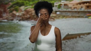 Sahilde, arka planda duran, burnuna dokunan, güneşli gökyüzünün altında beyaz atlet giyen bir kadın açık havada yaz havasının altını çiziyor..