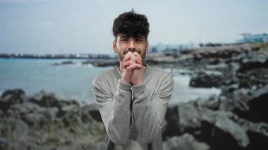 Elleri kumsalda sıkıca bağlı genç adam bulutlu gökyüzünün altında kayalık deniz kenarı zeminine karşı yalvarıyor..