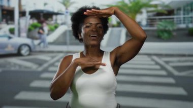 Kadın gülümsüyor ve şehir caddesinde kolsuz gömlekle işaret dili kullanıyor. Arka planda palmiye ağaçları ve arabalar var. Dışarıdaki sakin havayı yansıtıyor..