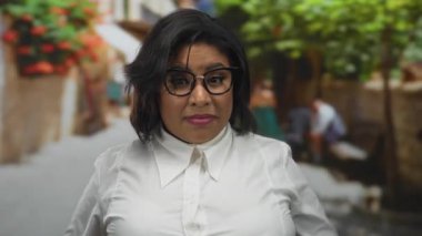 Gözlüklü ve beyaz tişörtlü kadın, canlı bir şehir caddesinde, canlı yeşillik ve mimariyle çevrili, kendinden emin bir ifade sergiliyor..