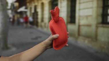 Şehrin caddesinde elinde kırmızı sıcak su şişesi tutan bir adam, el ve canlı kentsel geçmişin yakın plan detaylarını gösteriyor..