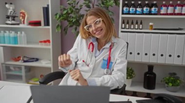 Gülümseyen kadın doktor, modern tıp ortamında steteskop ve bilgisayarla neşe ve profesyonelliği gösteren parlak klinik ofisinde kutlama yapıyor..