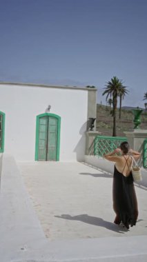 Lanzarote kanarya adalarında, açık gökyüzünün altında yeşil kapıları ve palmiye ağaçları olan beyaz bir binanın yanında gezen bir kadın.