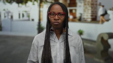 Şehir sokaklarında azim gösteren bir kadın, sıkılmış yumruğuyla güçlü bir poz veriyor ve kırmızı gözlüğü ve örgülü saçlarıyla ciddi bir ifade sergiliyor..