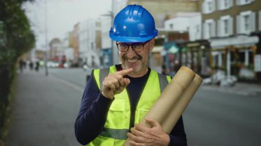 Mavi miğferli ve yüksek görüşlü yelek giyen olgun bir adam şehir merkezine öpücük gönderirken şehir sokaklarında planlar tutuyor..