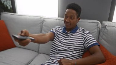 Kanepede uzanan genç adam elinde kumandayla rahat bir oturma odasında çizgili gömlek ve turuncu yastıkla oturuyor..