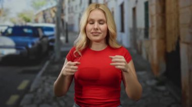 Sarışın genç bir kadın şehir sokaklarında jest yapıyor. Arka planda klasik arabalar olan kırmızı bir gömlek giyiyor. Duygularını ve ifadelerini aktarıyor..