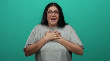 Gözlüklü ve uzun saçlı bir kadın, parlak yeşil bir arka plana karşı şaşkınlığını dile getiriyor, gündelik tarzda giyiniyor ve çevreden soyutlanmış, kıvrımlı figürler sergiliyor..