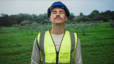 Sert şapkalı ve yansıtıcı yelek giyen genç İspanyol bir adam sakin bir parkta gözleri kapalı, yemyeşil ortamda sükuneti kucaklıyor, barışçıl farkındalığı sembolize ediyor..