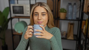 Modern ofiste oturan, kupa sarısı saçları olan yeşil kazak giyen, etrafı bitki ve raflarla çevrili bir kadın rahat bir iş ortamı yaratıyor.