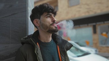 Kentsel sokak ortamında sigara içen genç adam, düşünceli bir ifade sergiliyor, sıradan bir kışlık ceket giyiyor ve arka planda arabalar ve binalar olan bir süveter giyiyor..