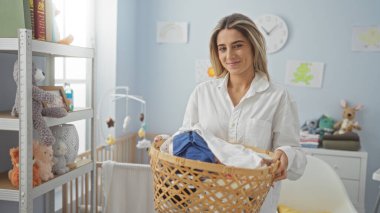 Çamaşır odasında gülümseyen bir kadın sepetini parlak bir iç mekânda tutuyor. Beyaz dekorlu, düzenli rafları ve doğal ışığı var. Huzur ve özeni özenle hazırlıyor..