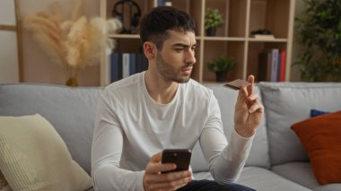 Genç İspanyol bir adam elinde telefon ve kredi kartıyla modern bir apartman dairesinde oturmuş online alışveriş veya finansal planlama tasvir ediyor..