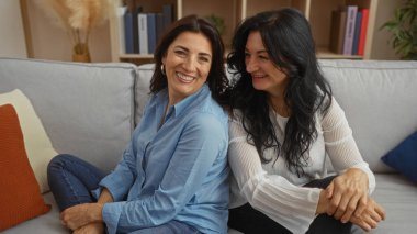 Sıcak bir oturma odasında birlikte oturan gülümseyen kadınlar, sıcaklığı ve aile bağlarını somutlaştıran, rahat bir ev ortamında olgun dostluğu ve mutluluğu gösteren..