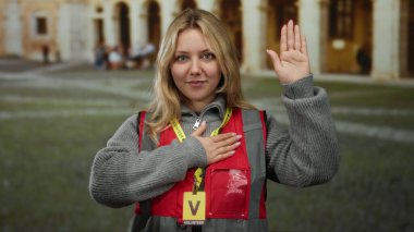 Açık hava üniversite ortamında, kırmızı yelek ve sarı saç giyen, eğitim ortamında kendini adamış ve bağlılık yemini eden bir kadın..