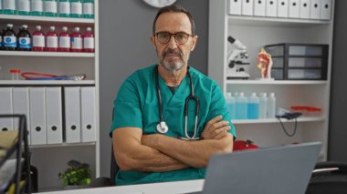 Gözlük ve deniz mavisi önlük takan İspanyol erkek doktor kolları çapraz bir hastane odasında güven içinde, tıbbi ekipman ve raflarla çevrili bir şekilde oturuyor..