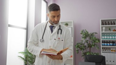Klinikteki doktor, profesyonel bir muayenehanede steteskop takmış, rafları ve bitkileri olan bir kitap okuyor..