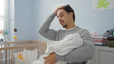 Yatak odasındaki genç adam battaniyeye sarılmış bebeği tutuyor. Parlak bir çocuk odasında beşiği ve şen şakrak dekorasyonları ile düşünceli görünüyor. Aile sevgisi ve yeni bir başlangıç olduğunu gösteriyor.