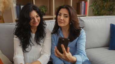 İki gülümseyen orta yaşlı kadın sıcak bir oturma odasında oturmuş, akıllı bir telefona bakarken birbirlerine eşlik ediyorlar, sıcak bir aile bağı sergiliyorlar..