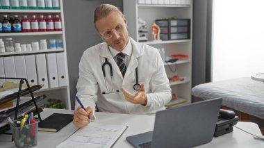 Klinikte, tıbbi aletler ve evrak işleriyle çevrili, profesyonel bir ortamda steteskop takan, bir hastanede video görüşmesi yapan sarışın beyaz bir adam..