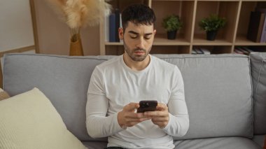 Genç İspanyol asıllı bir adam akıllı bir telefonla koltukta oturuyor. Etrafı modern ev dekorlarıyla çevrili. Rahat ve çağdaş bir yaşam tarzı sunuyor..