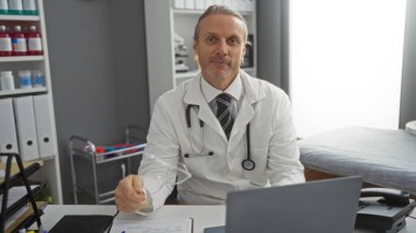 Klinikteki beyaz önlüklü, steteskoplu beyaz bir doktor profesyonel hastane ortamında kameraya bakıyor.