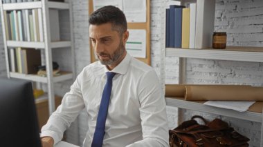 Modern bir ofiste çalışan, bilgisayara odaklanmış, resmi kıyafetler giyen, kitaplarla ve raflarla çevrili, profesyonel bir iş ortamını ima eden bir adam..