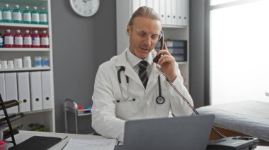 Hastane ofisinde çalışan doktor, tıbbi dosyalar ve raflarla çevrili telefon ve dizüstü bilgisayarla profesyonel bir sağlık ortamını resmediyor..