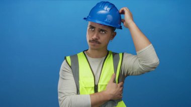 Yansıtıcı yelek ve kasketli genç İspanyol adam, izole edilmiş mavi duvar arkaplanına karşı düşünceli bir şekilde duruyor. İnşaatı ve planlamayı simgeliyor..