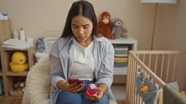 Bebek odasında, yumuşak ışıklı odada bebek eşyaları taşıyan bir kadın cep telefonu kullanıyor. Şefkat ve bağlı modern ebeveynlik gibi..