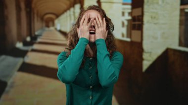 Üniversite koridorunda gülümseyen, neşeli duygular ve etkileyici jestler sergileyen bir kadın eski akademik ortamda yeşil bir kazak giyerken tuğla detaylarıyla.