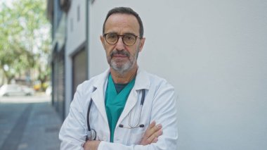 Beyaz önlüklü, gözlüklü ve sakallı kıdemli İspanyol bir adam bir şehir kliniğinin önünde güpegündüz kollarını kavuşturmuş duruyor..