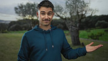 Serin bir park ortamında avucunun içini gösterip el hareketi yapan genç İspanyol bir adam..