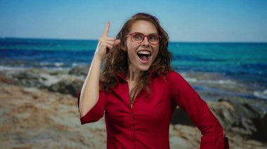 Kırmızı gömlekli kadın deniz kenarında neşeyle gülüyor, açık mavi deniz arka planına işaret ediyor, kumsaldaki mutluluk ve heyecanı somutlaştırıyor..