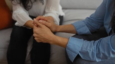 Konforlu bir oturma odasında el ele tutuşan iki kadın orta yaş bağını, aile sevgisini ve içeride arkadaşlığı yansıtıyor..