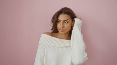 Beyaz süveter giyen kadın pembe arka planda poz veriyor, güzel ve esmer saçlarını ön plana çıkarıyor, gündelik ama zarif bir moda tarzı sergiliyor..