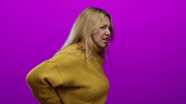 Sarışın kadın, parlak pembe bir duvarın önünde rahat sarı bir kazak giyerek sırt ağrısı çekiyor. Rahatsızlığını dile getiriyor ve izole bir ortamda sağlık ve yaşam tarzı temalarını yakalıyor..