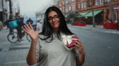 Yoğun şehir caddesinde elinde kalp şeklinde hediye kutusu olan bir kadın. Arka planda şehir yaşam tarzını gösteren bisikletçiler var..